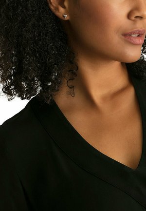 Black v-neck garment with a smooth texture, paired with small silver heart-shaped earrings. Features a natural curly hairstyle.