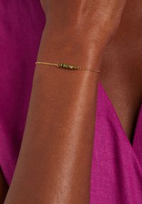 Pulsera de cadena de oro con cuentas verdes y doradas, llevada en un brazo bronceado, sobre un fondo de tela púrpura.