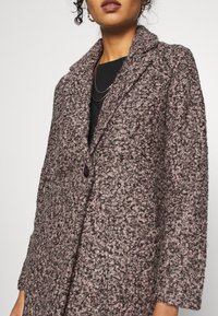 Femme portant un manteau texturé à motif bouclé mauve et noir à un seul bouton, par-dessus un haut noir avec des chaînes dorées superposées.