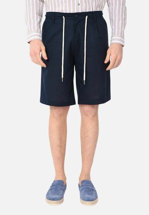 Uomo che indossa pantaloncini blu navy fino al ginocchio con cordini bianchi, camicia a righe con bottoni e mocassini blu, in piedi davanti a uno sfondo bianco.