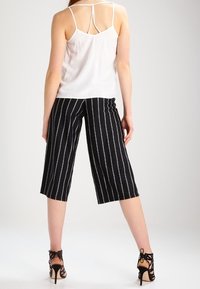 Mujer vistiendo un top blanco sin mangas y pantalones negros recortados con rayas verticales blancas, combinados con sandalias de tacón alto negras con cordones.