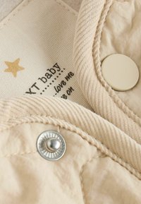 Beige Stoff mit gerippter Textur, versehen mit metallischen Druckknöpfen und einem Label mit gedrucktem Text sowie einem goldenen Stern-Detail.