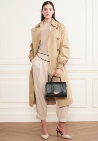 Manteau beige avec de grands boutons, léger pull, pantalon crème avec chaîne, sac à main noir texturé et talons crème. Textures lisses et polies.
