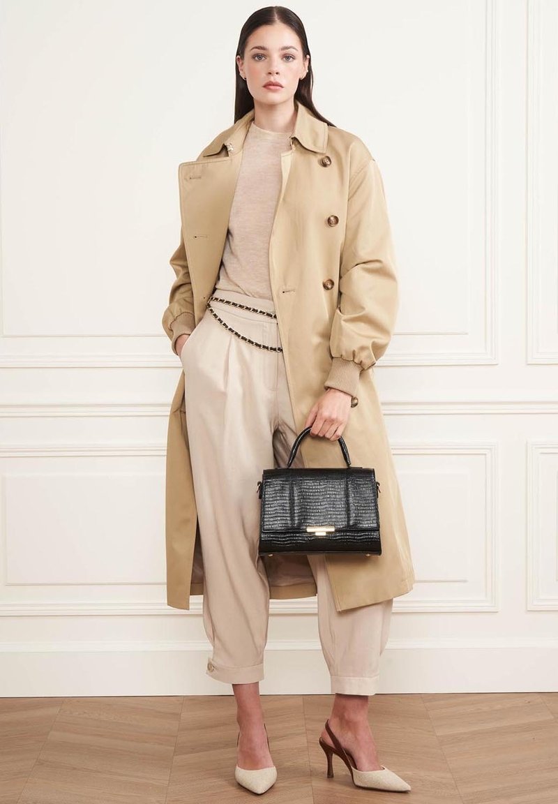 Manteau beige avec de grands boutons, léger pull, pantalon crème avec chaîne, sac à main noir texturé et talons crème. Textures lisses et polies.
