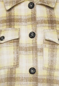 Flannel skjorte med et gult og brunt ternet mønster, sorte knapper og to brystlommer, lavet af blødt tekstureret stof.
