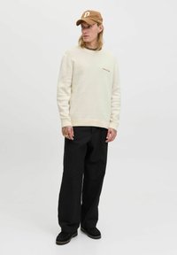 Cremfarbener Rundhalsausschnitt-Pullover mit gerippten Bündchen, kombiniert mit schwarzen weit geschnittenen Hosen und schwarzen Schuhen. Braune Kappe mit Logoakzent.