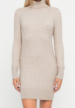 Femme portant une robe pull beige à manches longues, côtelée, avec un col roulé et une texture torsadée, debout devant un fond uni.