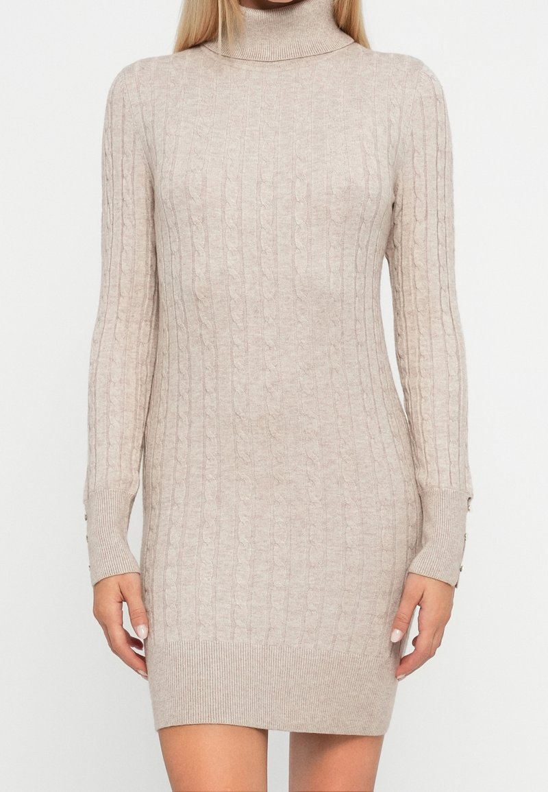 Femme portant une robe pull beige à manches longues, côtelée, avec un col roulé et une texture torsadée, debout devant un fond uni.