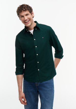 Young man smiling, wearing a dark green button-up shirt with rolled sleeves and blue jeans against a plain white background.