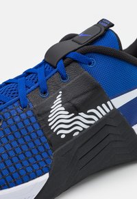 Modré a černé atletické boty s modrými tkaničkami, texturovanou boční částí a bílým vlnitým designem na černé střední části.