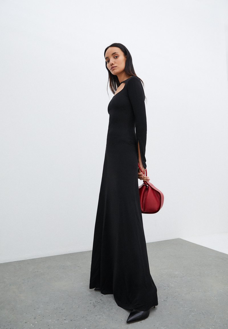 Vestido preto de mangas longas feito de tecido suave, com decote redondo e fendas laterais. Acessorizado com uma mala vermelha e sapatos pretos de bico.