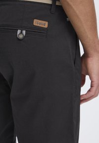 Pantalon noir en tissu léger, doté d'un label en cuir brun, d'une fermeture à bouton et d'une tirette rayée sur la poche arrière.
