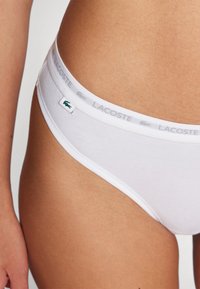 Culotte de bikini en coton blanc, avec une fine taille élastique ornée de l'inscription "LACOSTE" en gris et un petit logo crocodile vert.
