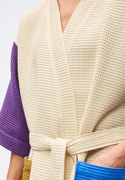 Άτομο που φοράει κρεμ ρόμπα με υφή βάφλας, με μανίκι σε μωβ χρώμα και τσέπες σε μπλε και μουσταρδί, δεμένη στη μέση με ζώνη.