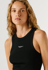 Femeie blondă cu păr creț, purtând un top negru fără mâneci cu logo-ul "Pepe Jeans" pe piept, expresie neutră, fundal simplu gri.