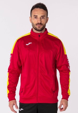 Chaqueta deportiva roja con detalles en amarillo, cierre completo, cuello alto y logo en el lado izquierdo del pecho. Presenta un diseño deportivo y una tela ligera.