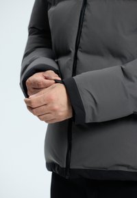 Puff-Jacke mit einer dunkelgrauen Außenschicht, Reißverschluss und schwarzen Manschetten. Eine Hand passt die Manschette an und zeigt eine glatte Textur.