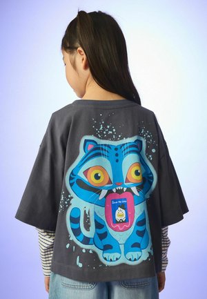 Dítě s dlouhými tmavými vlasy, které nosí tmavou nadměrnou košili s barevným modrým kresleným tygrem na zádech.