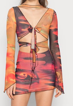 Femme portant une mini-robe à manches longues colorée avec des motifs abstraits orange, rouge et noir, et des découpes nouées au niveau de la poitrine et de la taille.
