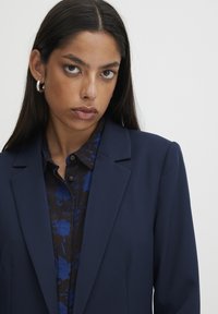 Blazer marine à revers structurés, associé à une chemise à motifs foncés avec des accents bleus. Le modèle porte des boucles d'oreilles en créole et a les cheveux raides.