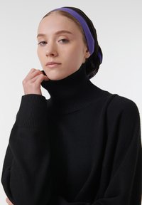 Zwarte coltrui met ribbeltextuur en hoge kraag, gedragen door een persoon met een meerkleurige hoofddoek tegen een neutrale achtergrond.