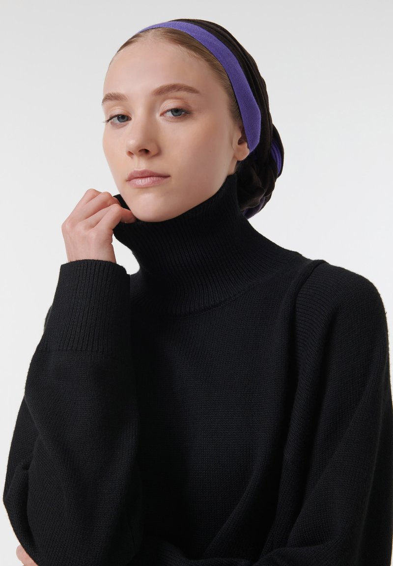 Zwarte coltrui met ribbeltextuur en hoge kraag, gedragen door een persoon met een meerkleurige hoofddoek tegen een neutrale achtergrond.