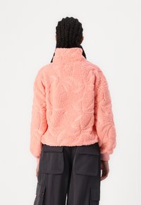 Vaaleanpunainen fleece-takki, jossa on korkea kaulus, teksturoitu kuvio ja ribbikäytävät. Päällä tummat cargo-housut, jotka osoittavat rentoa istuvuutta.