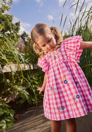 Noor tüdruk roosas ruudulises kleidis lillemustrilise tikandiga seisab õues kõrgete roheliste taimedega keskel, vaadates alla ja tasakaalu hoides sirutatud kätega.