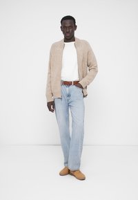 Beige knit cardigan with front zipper, white t-shirt, light blue jeans, and tan sneakers, standing against a plain white background.