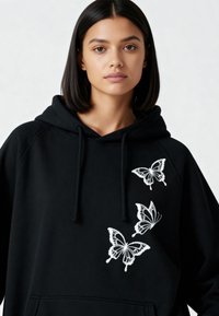 Sweat à capuche noir en tissu doux, avec trois papillons blancs brodés sur le devant. Cordons au niveau du cou et poche kangourou.