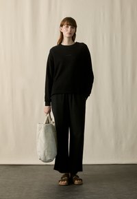 Sudadera negra con cuello redondo, combinada con pantalones negros holgados. Bolsa de tela ligera en gris claro y sandalias marrones con tiras visibles.