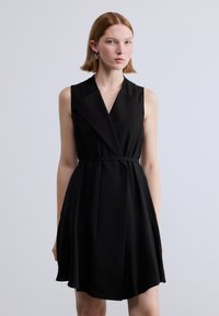 Μαύρο φόρεμα χωρίς μανίκια με σχέδιο τύλιγματος, V-λαιμόκοψη και ζώνη στη μέση. Διαθέτει λείο ύφασμα και ελαφρώς φουντωτή φούστα με διακριτικές πιέτες.