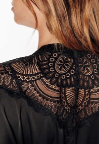 Vêtement noir présentant des détails en dentelle noire complexe au dos, mettant en valeur des motifs floraux et des bords festonnés sur un tissu lisse.