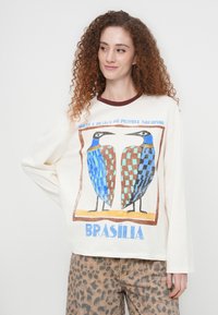 Κρεμ μπλούζα με μακριά μανίκια και μπορντό γιακά, διακοσμημένη με γραφικό δύο στυλιζαρισμένων πουλιών σε μπλε και καφέ χρώμα, με την επιγραφή "BRASÍLIA" από κάτω.