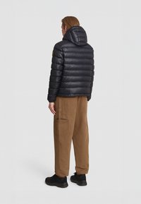 Giacca a vento nera con cappuccio, design a trapunte orizzontali e logo sulla spalla. Indossata con pantaloni cargo marroni e scarpe nere.