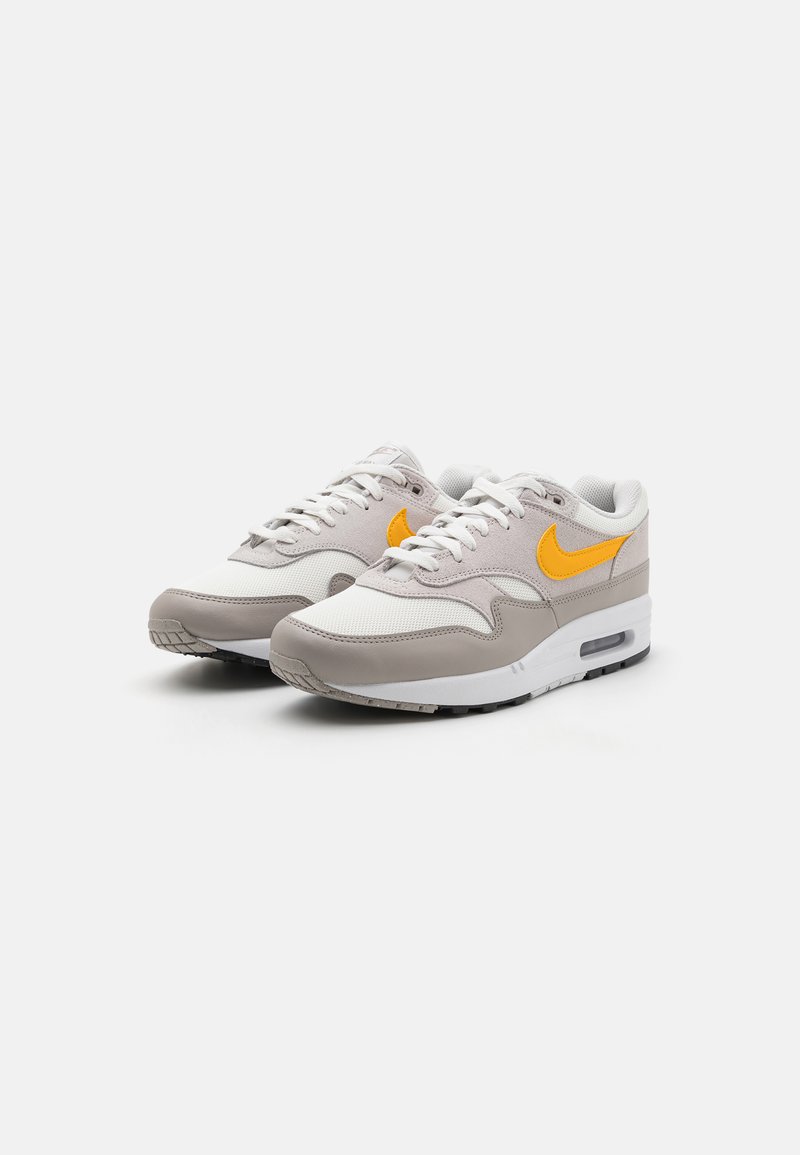 Zapatillas deportivas en gris y blanco con una parte superior de malla y cuero, que cuentan con un logo de Nike en color naranja, suela gruesa blanca y suela exterior texturizada negra.