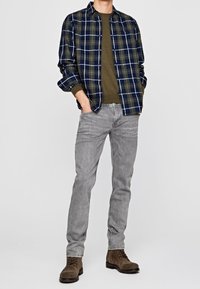 Camicia di flanella a bottoni in check blu navy e verde, t-shirt oliva, jeans slim-fit grigi e stivali marroni con lacci.