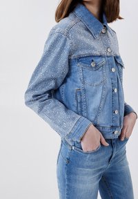 Nainen, joka on pukeutunut vaaleansiniseen farkkutakkiin, jossa on hopeiset niitit yläosassa, ja vastaaviin farkkuihin, kädet farkkujen taskuissa.