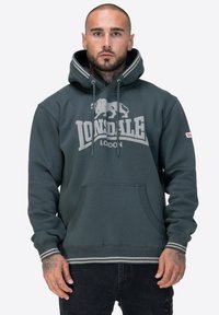 Sudadera de color verde oscuro con un bolsillo tipo canguro, acentos a rayas en contraste y un logo que presenta un león. Fabricada con material de felpa suave.