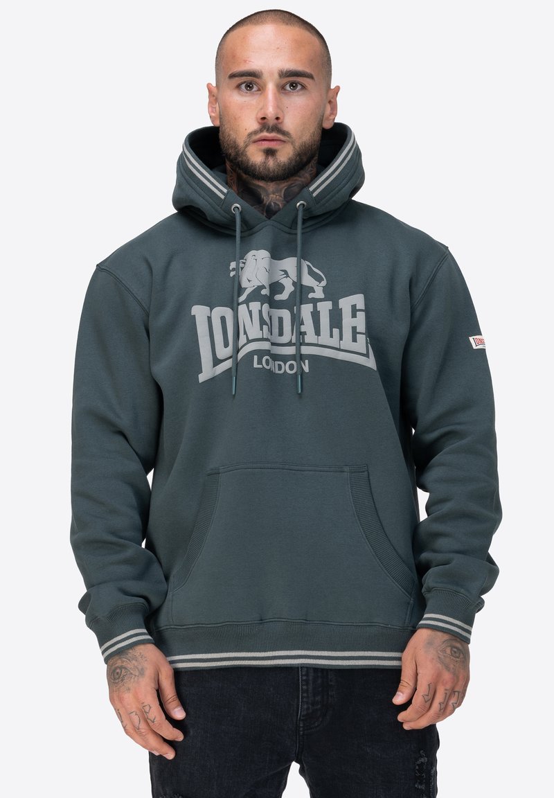 Sudadera de color verde oscuro con un bolsillo tipo canguro, acentos a rayas en contraste y un logo que presenta un león. Fabricada con material de felpa suave.