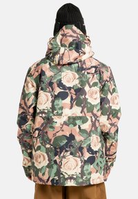 Osoba w kurtce z kapturem w kwiatowym wzorze moro, z różowymi różami i zielonymi liśćmi, zestawionej z brązowymi spodniami i czarną czapką beanie.