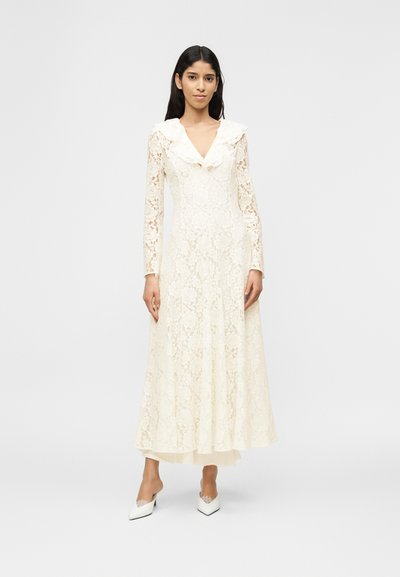 Γυναίκα που στέκεται, φοράει μακρυμάνικο, εκρού δαντελένιο φόρεμα με γιακά με βολάν και λευκά μυτερά τακούνια, μπροστά σε απλό φόντο.