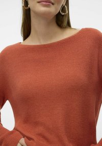 Femme portant un pull en tricot couleur rouille avec un large décolleté et de grandes boucles d'oreilles dorées, la main posée sur la taille.