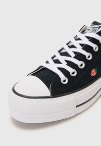 Baskets en toile noires avec un bout en caoutchouc blanc et une semelle blanche, dotées de lacets blancs et d'un petit détail en broderie de fraise sur le côté.