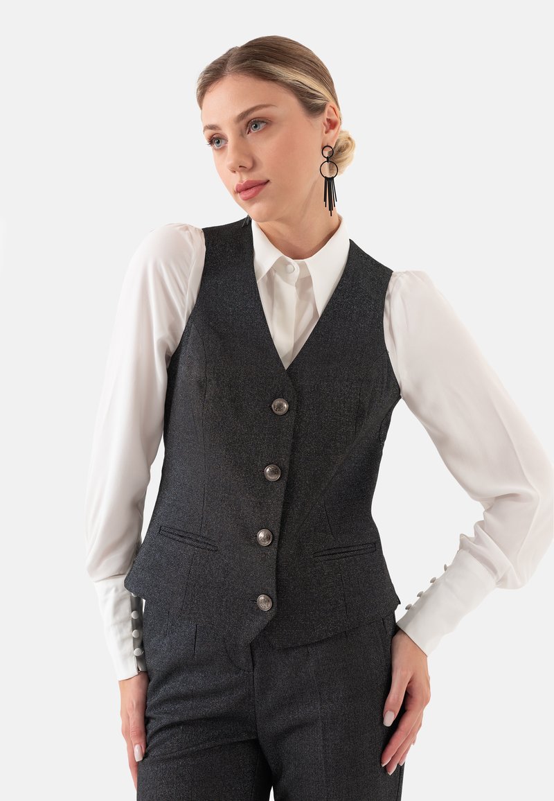 Donkergrijs op maat gemaakte gilet met een subtiele textuur, voorzien van een sluiting met zes knopen en twee voorkruiszakken. Onderneath een witte blouse met pofmouwen.