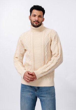 Suéter de cuello alto color crema con patrones de punto cable texturizados, con puños y dobladillo de canalé. Llevado con jeans de color azul claro.