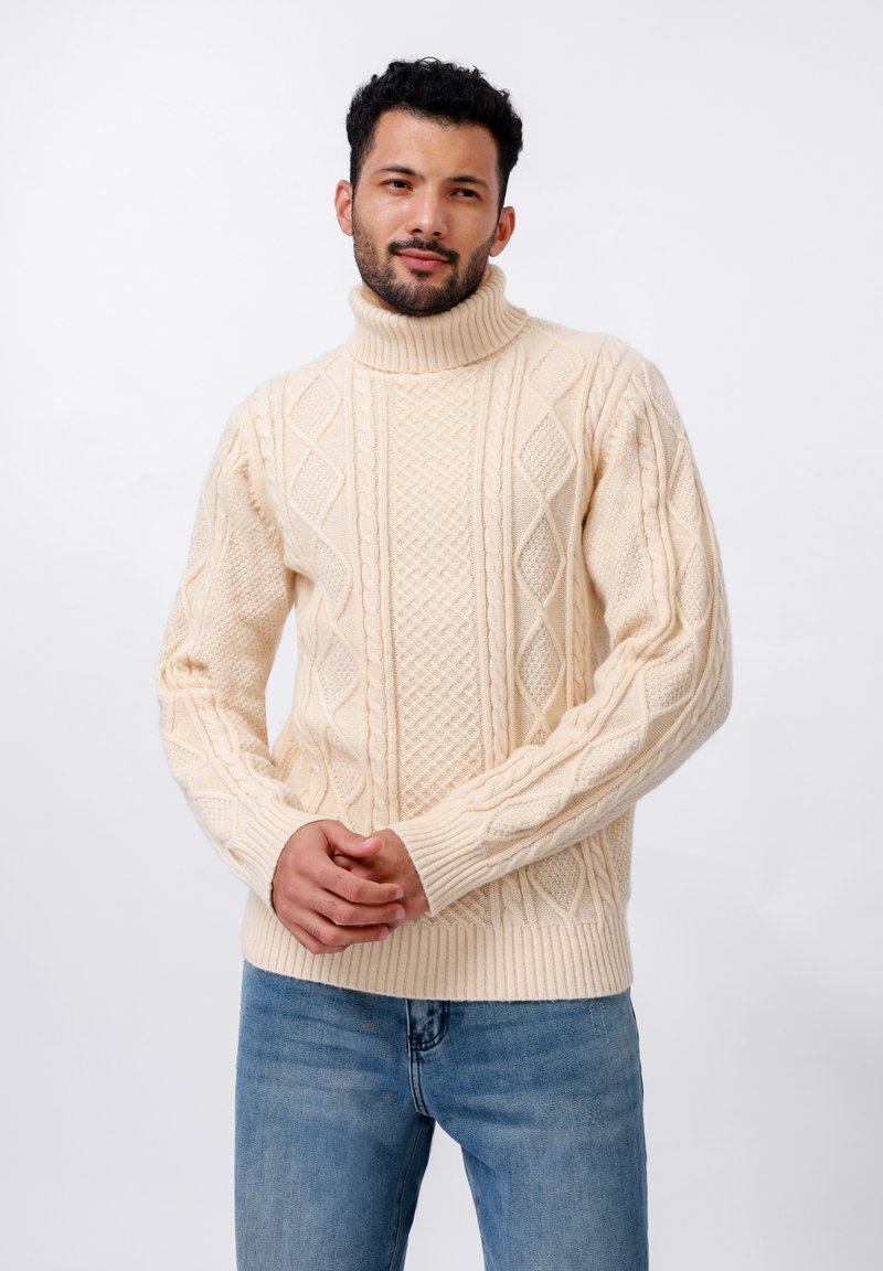 Pull en col roulé couleur crème avec des motifs de tricot en câble texturé, présentant des poignets et un ourlet côtelés. Porté avec un jean bleu clair.
