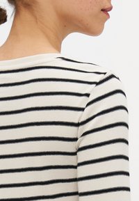 Stripet langermet topp med ribbet tekstur, med svarte striper på en kremfarget bakgrunn. Båtutringning og tettsittende silhuett.