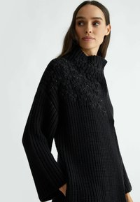 Černý pletený svetr s vysokým límcem, zdobený texturovanými květinovými vzory a korálkovými akcenty na ramenou, volnými rukávy a bočními kapsami.