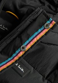 Suure lähedal must Paul Smith Juniori jakk, millel on mitmevärviline triibuline äär ja hõbedased nupud kaelusel.